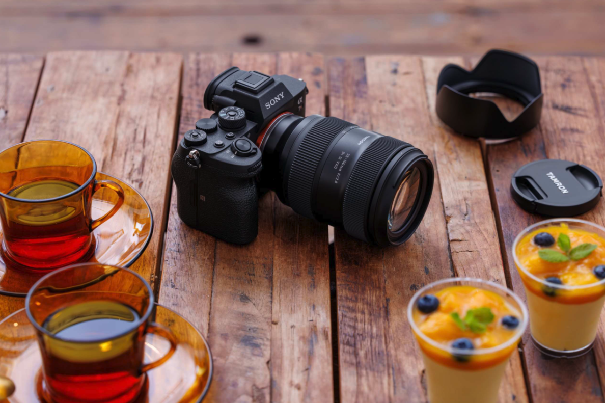Tamron camera with lens, glasses, and drinks on wooden table.