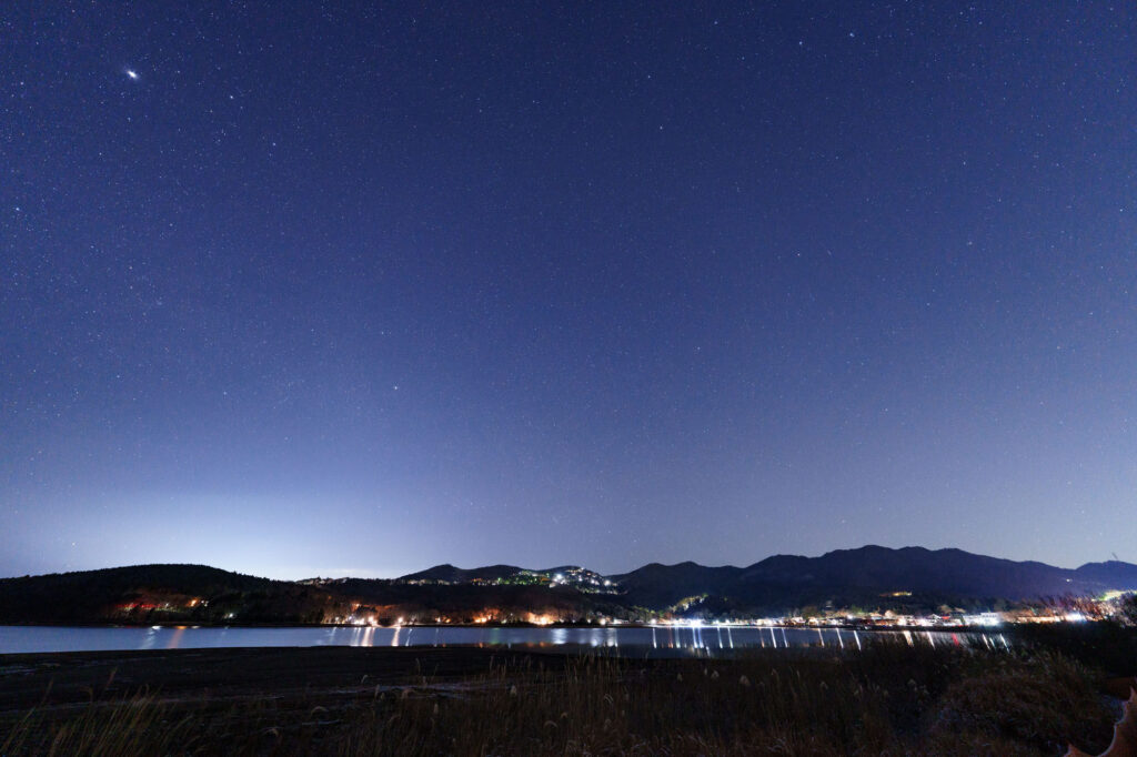 Night sky astrophotography with stars over a lake and city lights in the distance.